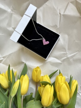 A pink heart-shaped pendant is displayed in a black jewelry box, surrounded by a bunch of yellow tulips. The jewelry box has a white exterior and is placed on a crumpled beige surface.