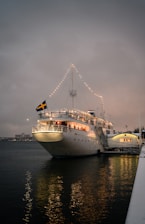 A large white ship is docked at the harbor, adorned with string lights along its top deck. The ship, named 'Lady Hutton Stockholm,' displays a Swedish flag at the stern. The calm water below reflects the lights, contributing to a serene atmosphere during dusk, as the sky appears overcast.