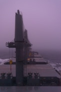 A large cargo ship is navigating through foggy weather on the ocean. The ship's deck is visible, featuring massive cranes and equipment. The atmosphere is misty and gray, with low visibility making the horizon indistinct.