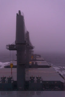 Cargo ship loaded with containers navigating through misty ocean waters under a fading sunset.