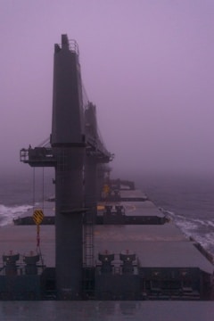A large cargo ship is navigating through foggy weather on the ocean. The ship's deck is visible, featuring massive cranes and equipment. The atmosphere is misty and gray, with low visibility making the horizon indistinct.