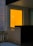 A small balcony or patio area with a wooden railing and a small wicker furniture piece. The focal point is a bright orange glass panel with a small round light source visible through it, suggesting either an interior light or the setting sun.