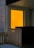A small balcony or patio area with a wooden railing and a small wicker furniture piece. The focal point is a bright orange glass panel with a small round light source visible through it, suggesting either an interior light or the setting sun.