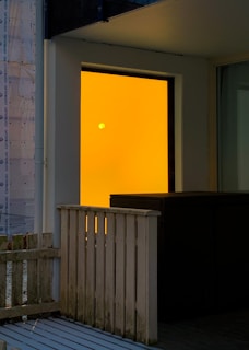 A small balcony or patio area with a wooden railing and a small wicker furniture piece. The focal point is a bright orange glass panel with a small round light source visible through it, suggesting either an interior light or the setting sun.