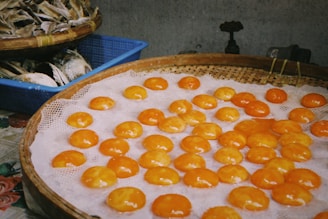A selection of bright orange salted egg yolks are neatly arranged on a woven bamboo tray, placed over a mesh. Nearby, in a blue plastic basket, there is a pile of dried fish. The setting suggests a local market atmosphere with a muted and rustic background.