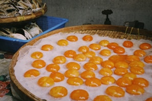A selection of bright orange salted egg yolks are neatly arranged on a woven bamboo tray, placed over a mesh. Nearby, in a blue plastic basket, there is a pile of dried fish. The setting suggests a local market atmosphere with a muted and rustic background.