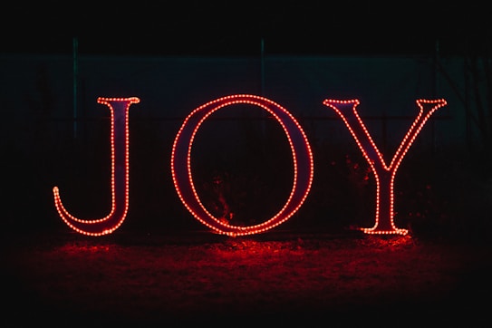Large letters spelling 'JOY' are illuminated in bright red lights against a dark background. The letters are made up of small, individual light bulbs, casting a warm glow on the surrounding area.