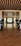 A luxurious hallway with a contemporary design, featuring sleek glass doors and large potted plants on both sides. Beyond the doors, a bright room with large windows allowing natural light in, decorated with curtains and several decorative elements including chairs and floral arrangements.