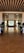 A luxurious hallway with a contemporary design, featuring sleek glass doors and large potted plants on both sides. Beyond the doors, a bright room with large windows allowing natural light in, decorated with curtains and several decorative elements including chairs and floral arrangements.