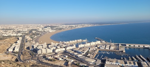 A coastal cityscape featuring a curving sandy beach along a blue ocean. The city buildings extend from the shore towards the background, with a mix of residential and commercial structures. A harbor with docks and various boats is present in the foreground, surrounded by infrastructure and roads. The landscape is expansive with clear skies overhead.