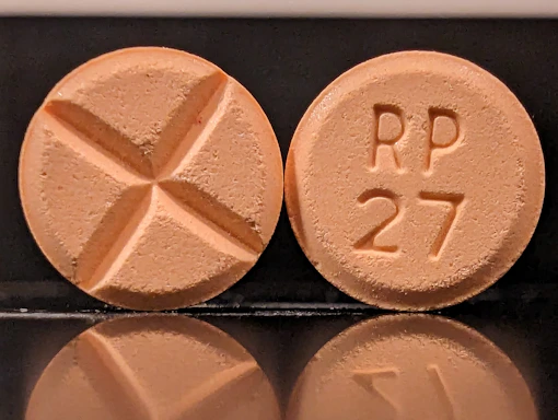 Two round tablets are displayed against a reflective black surface. One tablet features an engraved cross, while the other is marked with 'RP 27'. Both tablets have a consistent light orange color.