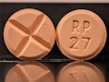 Two round tablets are displayed against a reflective black surface. One tablet features an engraved cross, while the other is marked with 'RP 27'. Both tablets have a consistent light orange color.