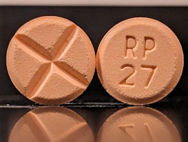 Two round tablets are displayed against a reflective black surface. One tablet features an engraved cross, while the other is marked with 'RP 27'. Both tablets have a consistent light orange color.
