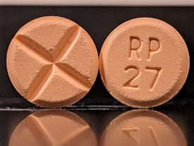 Two round tablets are displayed against a reflective black surface. One tablet features an engraved cross, while the other is marked with 'RP 27'. Both tablets have a consistent light orange color.