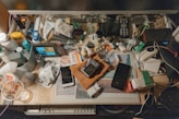 A cluttered desk is filled with a variety of everyday items including a monitor, remote control, game controller, smartphone, and various bottles and containers. Cables and electronic devices are scattered around, alongside papers and personal care products, creating a busy and chaotic impression.