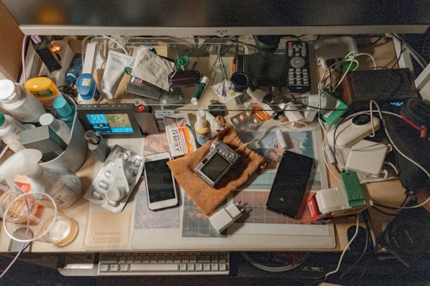 A cluttered desk is filled with a variety of everyday items including a monitor, remote control, game controller, smartphone, and various bottles and containers. Cables and electronic devices are scattered around, alongside papers and personal care products, creating a busy and chaotic impression.