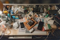 A cluttered desk is filled with a variety of everyday items including a monitor, remote control, game controller, smartphone, and various bottles and containers. Cables and electronic devices are scattered around, alongside papers and personal care products, creating a busy and chaotic impression.