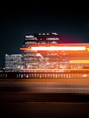 A nighttime cityscape featuring a brightly lit office building with the name 'asurion' visible. In front of the building, light trails create dynamic streaks of red, white, and orange, suggesting the movement of vehicles on a road or bridge. The surrounding area is dimly lit, emphasizing the illuminated structure.