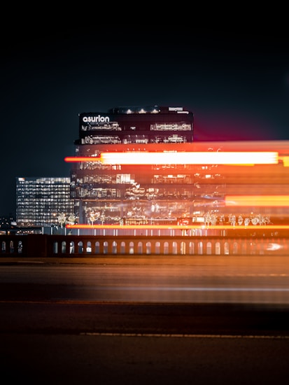 A nighttime cityscape featuring a brightly lit office building with the name 'asurion' visible. In front of the building, light trails create dynamic streaks of red, white, and orange, suggesting the movement of vehicles on a road or bridge. The surrounding area is dimly lit, emphasizing the illuminated structure.