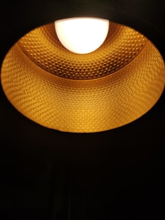 A close-up view of a textured golden lampshade with a bright white light bulb at the top center. The interior surface of the lampshade has a dotted pattern, creating a warm, glowing effect.
