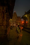 A person in traditional attire walks through the ancient stone corridor of a temple, illuminated by warm lights. The intricate architecture and carvings of the temple walls line the path, leading towards a towering gopuram under the evening sky.