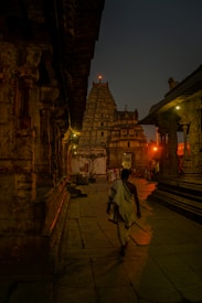 A person in traditional attire walks through the ancient stone corridor of a temple, illuminated by warm lights. The intricate architecture and carvings of the temple walls line the path, leading towards a towering gopuram under the evening sky.