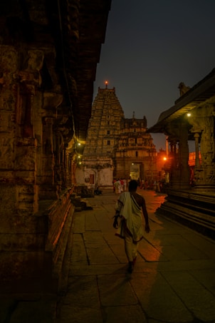 A person in traditional attire walks through the ancient stone corridor of a temple, illuminated by warm lights. The intricate architecture and carvings of the temple walls line the path, leading towards a towering gopuram under the evening sky.