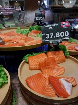 Slices of fresh salmon displayed on circular wooden trays, garnished with green lettuce. Price tags indicate the cost per 100g in Indonesian rupiah. The environment appears to be a supermarket or grocery store, with a focus on seafood.