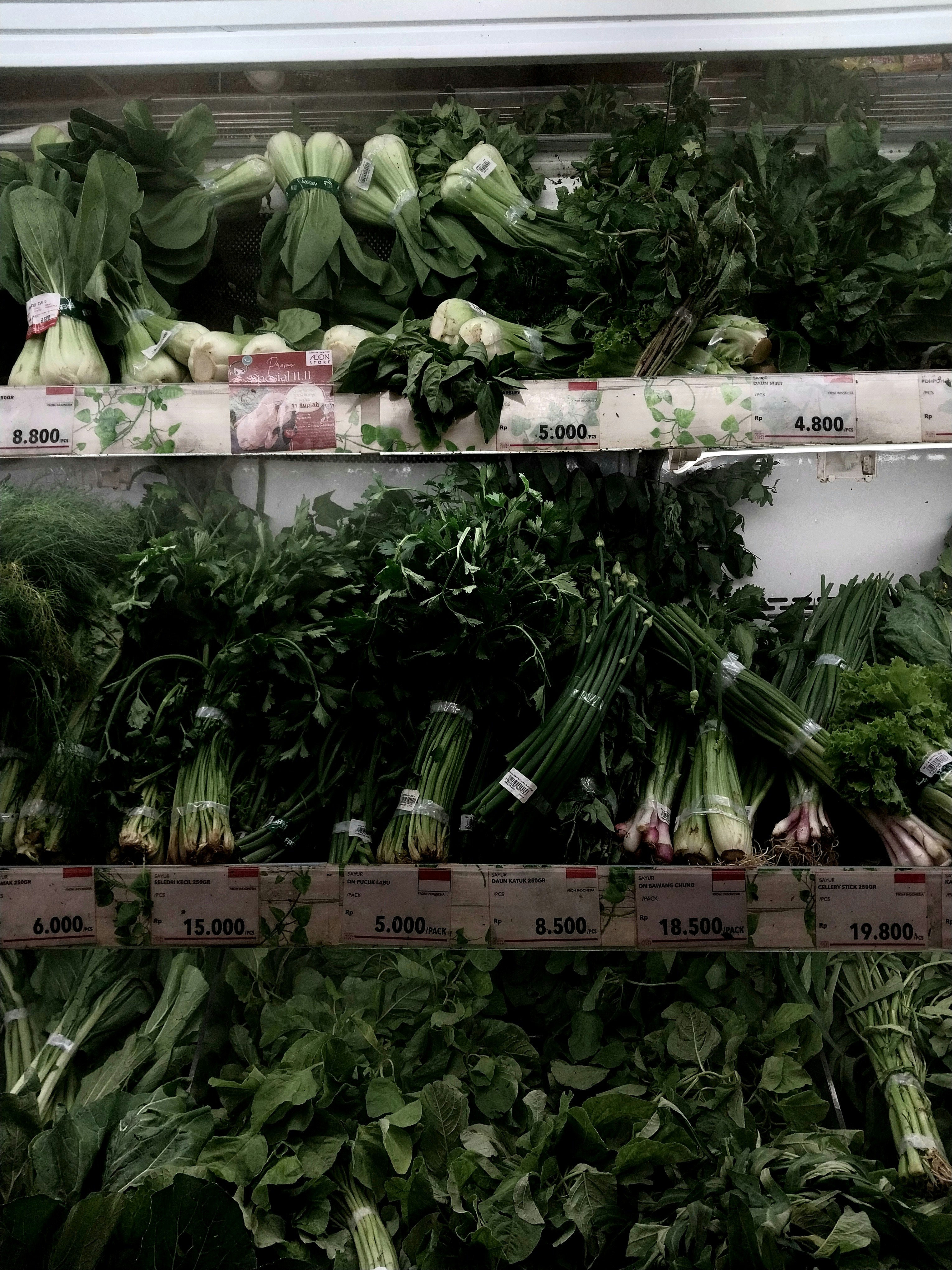 Fresh leafy greens and root vegetables bundled in clear plastic, arranged on shelves with price tags visible along the edge of the display.