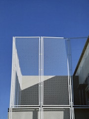 A tall metal fence with a chain-link design stands against a clear blue sky. The frame is white, and parts of a building are visible in the background, creating geometric shadows and shapes.