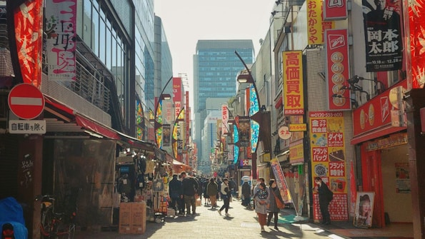 A bustling urban street scene with numerous people walking along a narrow path lined with shops. Bright, colorful signs and advertisements in various colors and languages adorn the buildings on both sides. Some stalls display goods near the sidewalk, creating a vibrant and busy atmosphere.