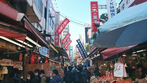 A bustling street market filled with numerous stalls on either side, colorful signs in Japanese hanging above. Many people are walking through the narrow lane, some browsing or buying goods. The atmosphere is lively, with visible interactions among the crowd.