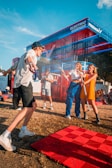 People are enjoying a lively event at an outdoor venue with a vibrant red and blue structure labeled 'Electric Corner' and 'Free Now' in the background. Attendees are interacting playfully, with some holding drinks and wearing casual summer attire. The atmosphere is energetic and fun under a clear blue sky.