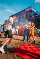 People are enjoying a lively event at an outdoor venue with a vibrant red and blue structure labeled 'Electric Corner' and 'Free Now' in the background. Attendees are interacting playfully, with some holding drinks and wearing casual summer attire. The atmosphere is energetic and fun under a clear blue sky.