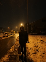 A young person wrapped in a thick coat, standing in softly falling snow under dim streetlights.