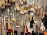 A variety of cleaning brushes hanging on a metal perforated wall. The brushes vary in shape, size, and bristle type, including long-handled brushes, round scrubbers, and small handheld brushes. The materials range from wooden handles to metal wire loops, and bristle colors are primarily beige, white, and brown.