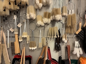 A variety of cleaning brushes hanging on a metal perforated wall. The brushes vary in shape, size, and bristle type, including long-handled brushes, round scrubbers, and small handheld brushes. The materials range from wooden handles to metal wire loops, and bristle colors are primarily beige, white, and brown.
