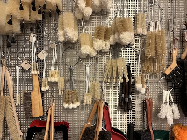 A variety of cleaning brushes hanging on a metal perforated wall. The brushes vary in shape, size, and bristle type, including long-handled brushes, round scrubbers, and small handheld brushes. The materials range from wooden handles to metal wire loops, and bristle colors are primarily beige, white, and brown.