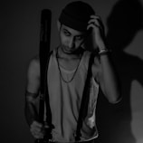 A young man is standing in a dimly lit room, wearing a sleeveless shirt, suspenders, and a beanie. He has a serious expression, holding a baseball bat in one hand, casting a prominent shadow on the wall behind him.