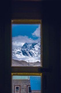 A majestic mountain peak covered in snow is visible through a window. The view is framed by the dark outline of the window, providing a stark contrast to the bright blue sky and white clouds surrounding the mountain. Below the mountain, a building with a green roof and stone walls is partially visible, adding an element of human presence to the scene.