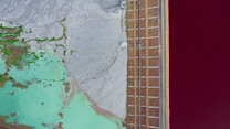 An aerial view captures a striking landscape divided into distinct sections. On the left, a mix of turquoise water and green vegetation contrasts sharply with an adjacent gray, barren area. To the right, a geometric pattern of brown, grid-like structures runs parallel to a vibrant red expanse, possibly a body of water.