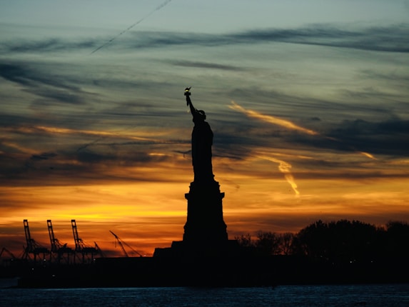 A silhouette of the Statue of Liberty against a vibrant sunset with an array of colors, including deep orange and yellow, spread across the sky. The skyline includes industrial cranes, adding to the urban landscape. The water in the foreground reflects the evening tones.