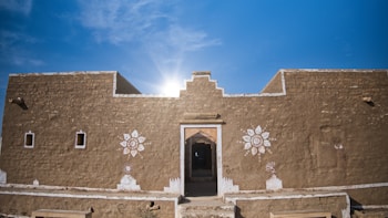 A rustic clay building with intricate white floral designs painted on its exterior, prominently featuring a central doorway. The architecture appears simple yet traditional, with a flat roof under a clear blue sky with the sun glowing behind the structure.