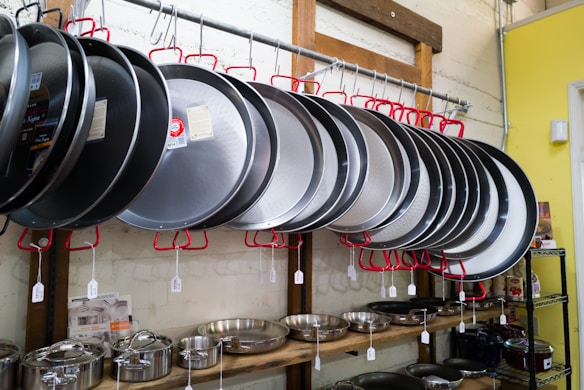 A selection of large, silver cooking pans hangs on red hooks from a metal rod in a store. Below them, various pots and pans are displayed on wooden shelves against a light-colored wall.