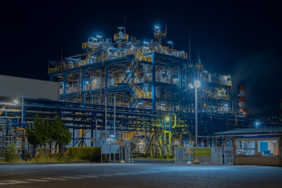 A large industrial complex is illuminated at night, with an intricate network of pipes and structures. Bright, artificial lighting highlights the metal framework, creating a contrast against the dark sky. Trees and hedges line the perimeter, while a building with a fenced-off area is visible in the foreground.