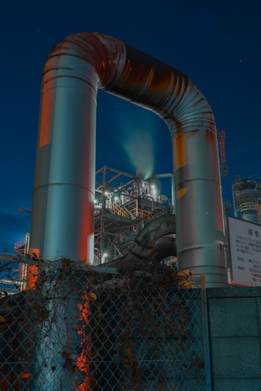 A large, curved metal pipe structure stands prominently against a backdrop of industrial machinery and scaffolding. The setting appears to be a factory or industrial plant at night, with lights illuminating various metal components. In the foreground, there is a chain-link fence with overgrown vegetation.
