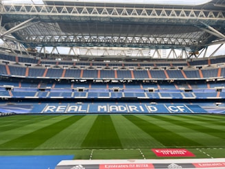 A large, empty football stadium with a well-maintained green field. The stands are primarily blue with white lettering spelling out 'REAL MADRID CF' across the seats. The roof structure is visible, showcasing metal beams and supports. There are some advertisements along the boundary of the pitch, including one from Emirates.