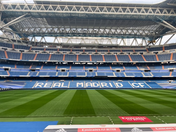 A large, empty football stadium with a well-maintained green field. The stands are primarily blue with white lettering spelling out 'REAL MADRID CF' across the seats. The roof structure is visible, showcasing metal beams and supports. There are some advertisements along the boundary of the pitch, including one from Emirates.
