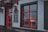 A storefront with red accents featuring a door and window. A chalkboard sign is displayed beside the door with writing in another language. Through the window, an illuminated decoration resembling a deer is visible. The scene is dimly lit, suggesting overcast weather.