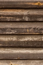 A series of horizontal, rustic wooden logs with visible texture and natural weathering. The wood is aged, with rough surfaces and varying shades of brown, interspersed with small, natural imperfections.